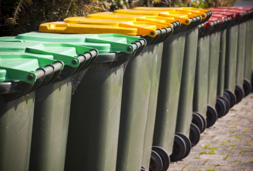 A row of large outdoor wheelie bins positioned on a paved surface, aligned alongside each other and extending into the background. The bins have matching dark grey bases with smooth, slightly reflective surfaces. The lids are brightly coloured in green, yellow, and red, with the green lids in the foreground, followed by yellow and red ones further back. The lids are made of plastic with a textured surface and are fitted with black hinges. The bins are arranged on a concrete pavement with a few small patches of moss or grass growing between the cracks. The background shows blurred greenery, suggesting the bins are located near a residential or commercial building in Rotherhithe, with natural daylight illuminating the scene. This setup is typical for waste management and rubbish collection services, exemplified by companies like Office Clearance Rotherhithe, servicing local rubbish removal needs.