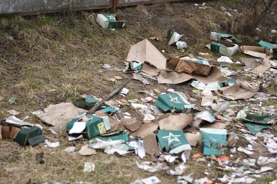 An aerial view displaying a large accumulation of mixed waste and debris scattered across a patch of land adjacent to a gravel pathway and a grassy area with sparse trees. The waste includes plastic b