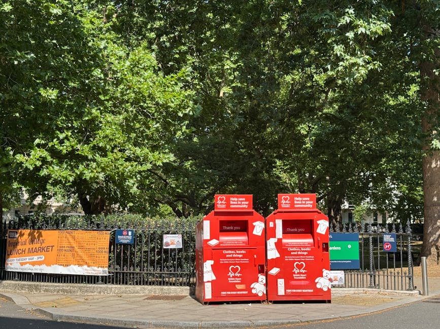 Two red charity donation bins are positioned on a paved sidewalk in front of a black metal fence, adjacent to a small green public garden area. The bins are rectangular with sloped tops, featuring whi