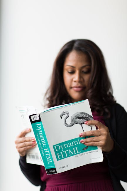 A woman with medium-dark skin and long dark hair is standing indoors against a plain white background, holding and reading a book titled 'Dynamic HTML' with a cover featuring a bird illustration, poss